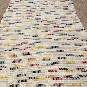 May include: Cream-colored rectangular rug with a woven texture. The rug features a pattern of horizontal stripes in red, yellow, navy, and black. The rug is made of a soft, durable material.