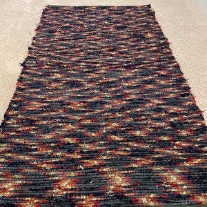 May include: A rectangular, multi-colored rug with a woven texture. The rug features a mix of dark blue, red, and tan threads, creating a textured pattern. The edges are finished with a green border. This rug is suitable for home decor.