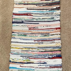 May include: A rectangular, woven rag rug with a striped pattern. The rug features a variety of colors, including white, red, blue, pink, and green. The rug is approximately 24 inches long and 12 inches wide.