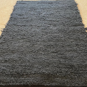 May include: A rectangular, dark gray rug with a textured, shaggy surface. The rug has a black border along the bottom edge and a slightly frayed edge on the top. The rug is made of a woven material.
