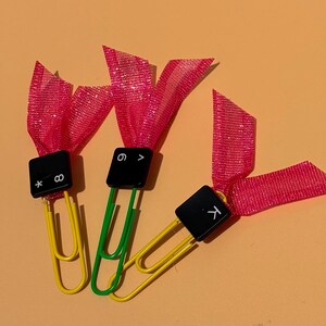May include: Three paper clips, two yellow and one green, each attached to a black square keyboard key cap. The keys are labeled with an asterisk and infinity symbol, the number six and a greater-than sign, and the letter K. Each has a pink ribbon.
