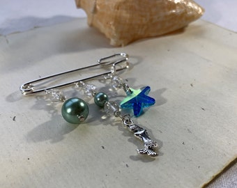 May include: A silver safety pin with a green pearl, a clear crystal, and a blue iridescent starfish charm. A silver mermaid charm hangs from the bottom of the pin.