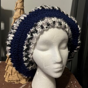 May include: A blue and white crocheted beanie with a black and white pattern. The beanie is on a white mannequin head.