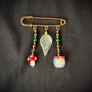 May include: A gold safety pin with three charms: a green leaf, a red and white mushroom, and a colorful owl. The charms are attached to the pin with black and green beads.