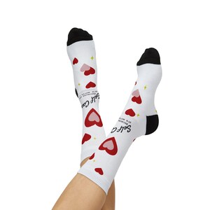 May include: White crew socks with black cuffs and toes, featuring a repeating pattern of red and pink hearts, small yellow four-pointed stars, and the black text "Self Care Because you are worth it" printed vertically along the side of each sock.
