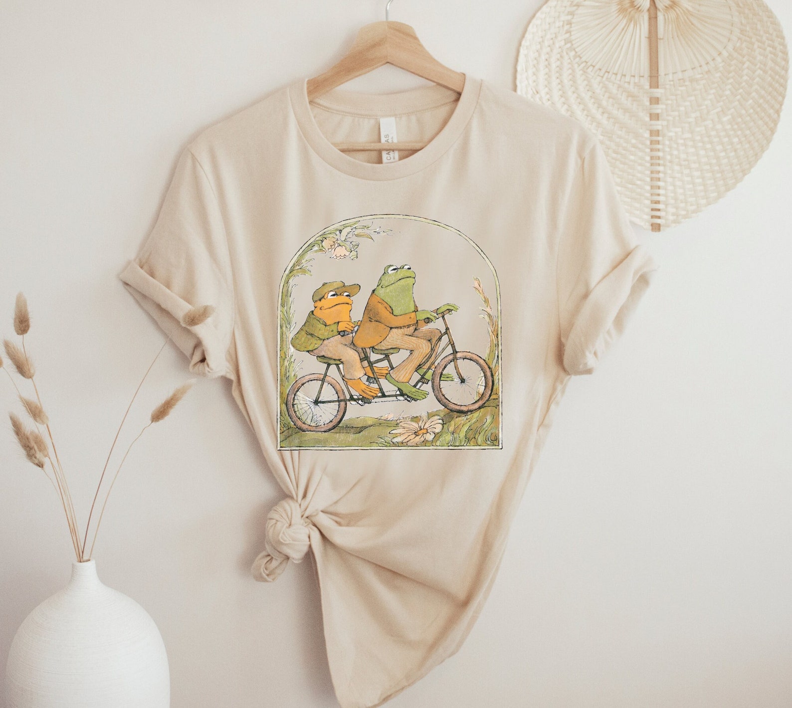 vanilla colored T-shirt with two frogs riding a bicycle