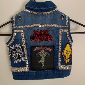 May include: A blue denim vest with silver studs and various band patches, including "Alice Cooper", "Motley Crue", and "Ghost".