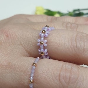 Puede incluir: Dos anillos delicados hechos de pequeñas cuentas moradas con detalles dorados. El anillo de arriba está hecho de cuentas con forma de flor, mientras que el anillo de abajo está hecho de cuentas redondas.