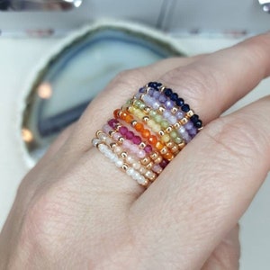Könnte beinhalten: Ein Stapel von 12 Ringen aus kleinen Edelsteinperlen in einem Regenbogen von Farben. Die Ringe werden an einem Finger getragen.