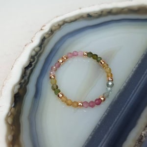 Könnte beinhalten: Ein mehrfarbiges Edelstein-Armband mit kleinen, runden Perlen in Rosatönen, Grün und Gelb. Das Armband ist aus goldfarbenen Metallperlen gefertigt und wird auf einer weißen und grauen Achatplatte präsentiert.