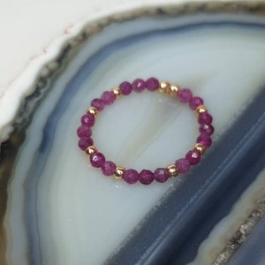 Op de afbeelding: Een delicate ring gemaakt van kleine, gefacetteerde, roze edelstenen en gouden kralen. De ring wordt getoond op een witte en grijze agaatplak.