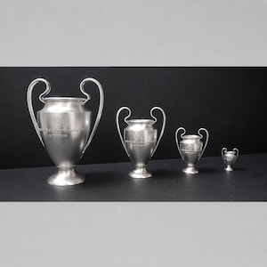 May include: Four silver-colored metal trophies arranged in descending order of size. Each trophy features two curved handles, a wide rim, and a pedestal base. The text "COUPE DES CLUBS CHAMPIONS EUROPÉENS" is engraved on the front of each trophy.
