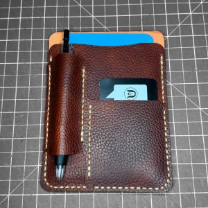 May include: A dark brown textured leather card holder with a vertical pen loop on the left side containing a black pen. The holder features a front pocket with a black card displaying a white speech bubble icon and a blue card tucked inside.