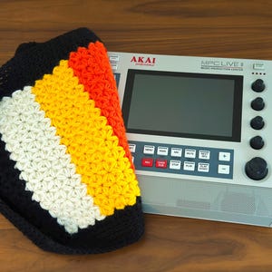 Peut inclure: Un chapeau crocheté noir avec un motif rayé coloré blanc, jaune et orange est posé à côté d'un centre de production musicale AKAI MPC Live II argenté. Le chapeau présente des rayures verticales. Le centre de production musicale a un grand écran et des boutons.