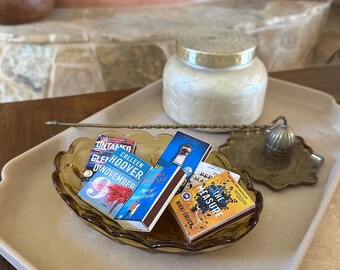 Könnte beinhalten: Ein dekoratives Tablett zeigt eine Sammlung von Gegenständen. Ein Glas mit goldenem Deckel, ein Messing-Räucherstäbchenhalter und eine kleine bernsteinfarbene Glasschale mit drei büchergroßen Streichholzschachteln. Die Bücher tragen die Titel "November 9", "Part of Your World" und "The Measure".