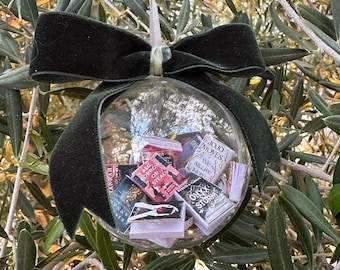May include: A clear glass Christmas ornament filled with miniature books, suspended by a green velvet ribbon and bow. The ornament is hanging from a tree branch with green leaves. The miniature books have titles like "Onyx Storm" and "Beg, Borrow or Steal".