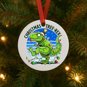 May include: A circular Christmas tree decoration featuring a green dinosaur wearing a Santa hat and festive lights. The text "CHRISTMAS TREE-REX" is displayed around the edge. A red ribbon is attached for hanging on the tree.