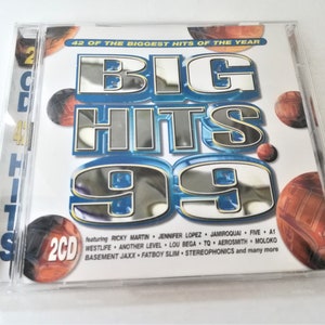 Puede incluir: Un conjunto de dos CD titulado "Big Hits 99" con 42 de los mayores éxitos del año. La portada presenta un diseño plateado y azul con el título en letras grandes y en negrita. El conjunto de CD incluye artistas como Ricky Martin, Jennifer Lopez, Jamiroquai, Five, A1, Westlife, Another Level, Lou Bega, y muchos más.