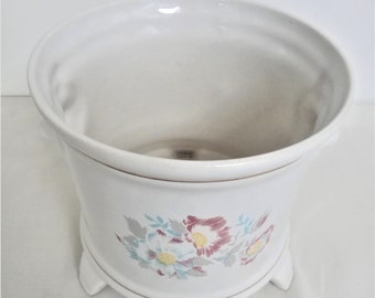 May include: White ceramic planter with a floral design featuring pink, yellow, and green flowers. The planter has three legs and a gold rim.