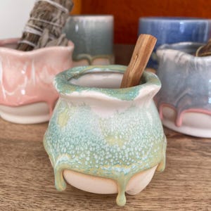 May include: A small, handmade ceramic pot with a light green and white speckled glaze, holding a piece of Palo Santo wood. The pot has a unique, dripped glaze design. Other similar pots in pink, blue, and teal are in the background.
