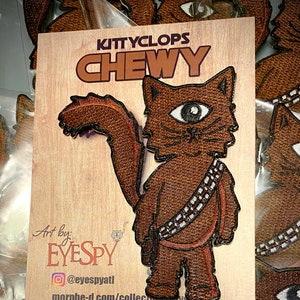May include: A brown embroidered patch of a cat with one eye, wearing a bandolier and a belt. The cat is standing on its hind legs. The text "KITTYCLOPS CHEWY" is at the top of the patch. The text "Art by: EYESPY @eyespyatl morphe-d.com/collections/eyespy" is below the patch. The text "TACTICOOL IMAGING tacticoolimaging.com" is at the bottom of the patch.