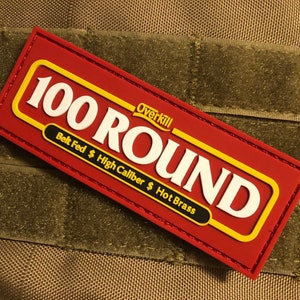 Pode incluir: Patch vermelho e amarelo de 100 Round com texto branco. O patch tem um verso de velcro e diz "Overkill" e "Belt Fed $ High Caliber $ Hot Brass".