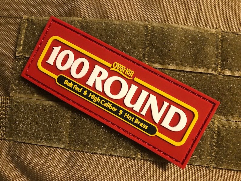 K&ouml;nnte beinhalten: Roter und gelber 100 Round Patch mit wei&szlig;em Text. Der Patch hat einen Klettverschluss und lautet "Overkill" und "Belt Fed $ High Caliber $ Hot Brass".