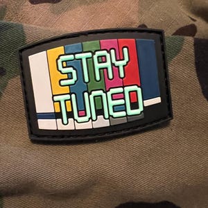 May include: Black and white patch with a colorful television screen design. The text "STAY TUNED" is written in green pixelated letters.