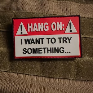 以下が含まれることがあります： 赤と白の背景に「HANG ON: I WANT TO TRY SOMETHING...」と白い文字で書かれた長方形のパッチ。 パッチは黒い縁取りがあり、質感のあるベージュ色の背景に配置されています。