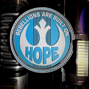 May include: A round, blue and white emblem with the text "REBELLIONS ARE BUILT ON HOPE". The emblem features a stylized bird symbol and is set against a dark background with metallic accents. The design is reminiscent of a popular science fiction theme.