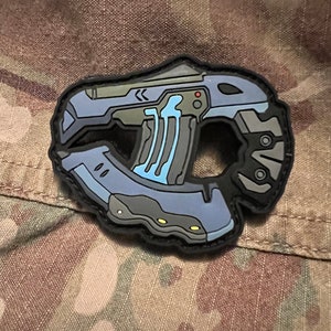 May include: A blue and black tactical patch featuring a futuristic pistol with a blue energy beam. The patch has a black border and a textured surface.