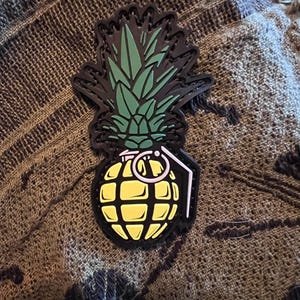 May include: A black, yellow, and green patch in the shape of a pineapple grenade. The top of the patch features green pineapple leaves, while the bottom is a yellow grenade with a white outline.