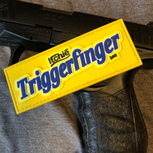 以下が含まれることがあります： 青と白で「Itchie Triggerfinger bar」の文字が入った黄色のパッチは、チョコレートバーに似ており、黒い拳銃の上に置かれています。 パッチには黄色の縁取りがあり、灰色の背景に配置されています。