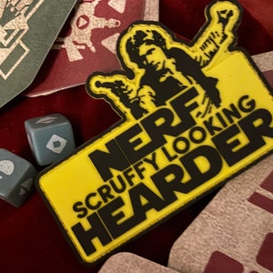 以下が含まれることがあります： 黄色と黒のパッチに、スターウォーズのハン・ソロの漫画の画像が描かれています。パッチのテキストは「NERF SCRUFFY LOOKING HEARDER」です。