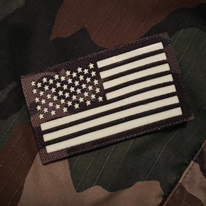 Peut inclure: Un patch de drapeau américain blanc avec 50 étoiles et 13 bandes sur un fond gris foncé. Le patch est cousu sur un tissu camouflage.