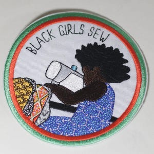 May include: A white embroidered patch with a green and orange border. The patch features a black girl sewing on a sewing machine with the text "BLACK GIRLS SEW" in black.