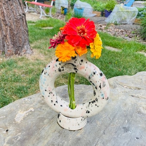 May include: A white ceramic vase with black and pink splatter paint. The vase is shaped like a ring and has a small base. It is filled with a bouquet of orange and red flowers.