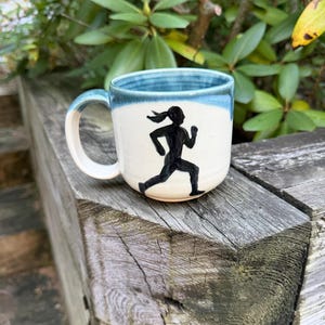 May include: A ceramic mug with a cream-colored body and a blue glazed rim. The mug features a black hand-painted silhouette of a person running, depicted in a dynamic stride with arms bent and hair flowing back, centered on the front surface.