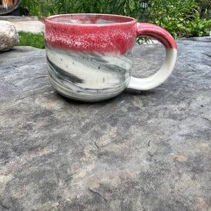 May include: A handmade ceramic mug with a red rim and handle, transitioning to white and gray marbled patterns on the body. The mug has a curved handle and a wide opening, perfect for coffee or tea.