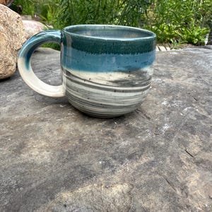 May include: A handmade ceramic mug with a rounded shape and a large handle. The mug features a color gradient of teal, blue, and gray, with a white handle. The mug is sitting on a gray stone surface.