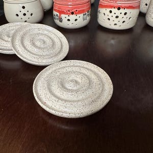 May include: A set of white ceramic garlic keepers with red lids and small holes. The lids have a speckled design. The keepers are sitting on a dark brown wooden surface with two white ceramic coasters.