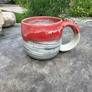 May include: A handmade ceramic mug with a red and white glazed rim and handle. The body of the mug features horizontal bands of gray and blue. The mug is designed for hot beverages and is perfect for coffee or tea.