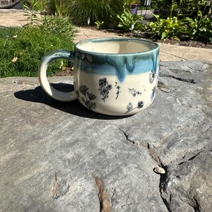 May include: A handmade ceramic mug with a cream-colored base and a teal blue rim. The mug features a floral design in black and white. The handle is also teal blue. The mug is sitting on a gray stone surface.