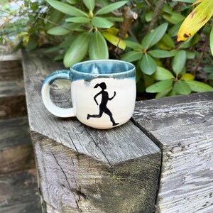 May include: A ceramic mug with a cream-colored body and a blue glazed rim. The mug features a black silhouette of a person running on the front. It has a curved handle on the left side and a smooth, rounded shape with a glossy finish on the upper rim.