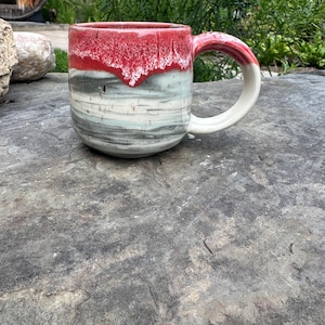 May include: A handmade ceramic mug with a red rim and handle, transitioning to a marbled gray, white, and blue body. The mug has a rounded shape and a white handle. The mug is sitting on a gray stone surface.