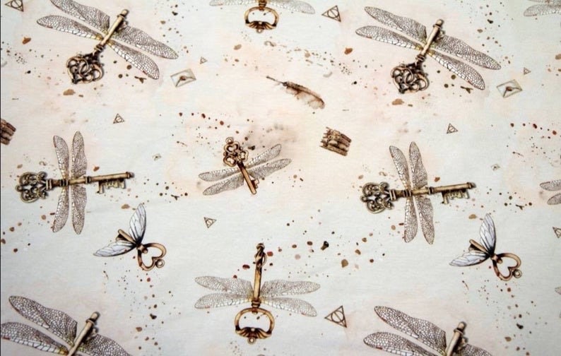 Puede incluir: Una tela blanca con un patr&oacute;n repetido de alas de lib&eacute;lula y llaves. Las llaves son ornamentadas y tienen un aspecto vintage. El fondo es beige claro con salpicaduras de pintura marr&oacute;n.