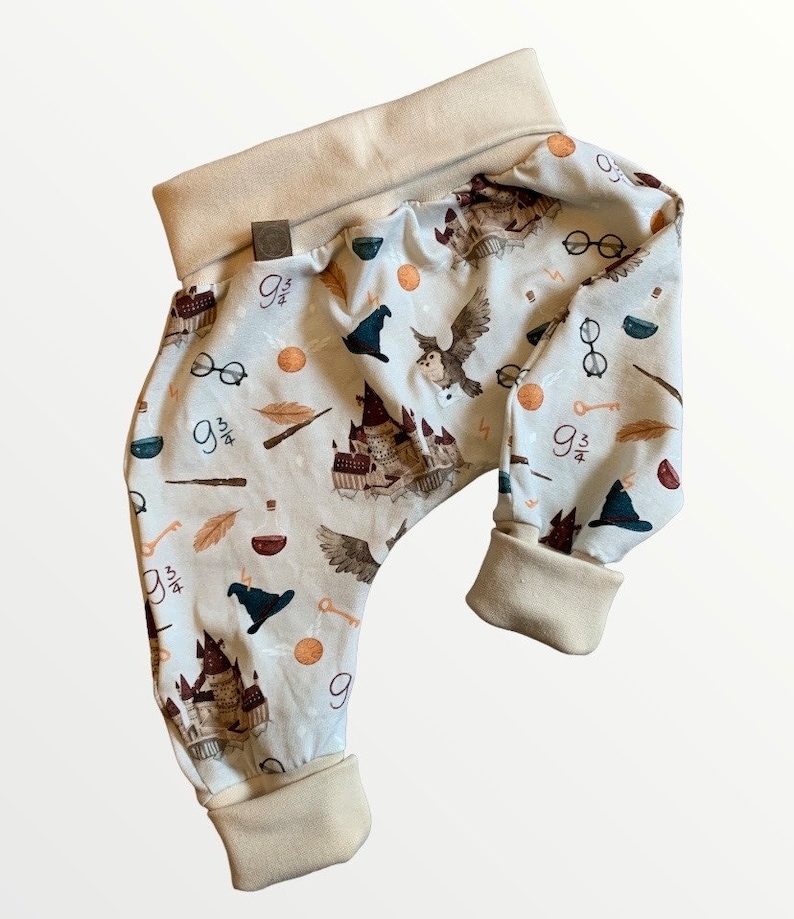 Puede incluir: Un pantal&oacute;n de beb&eacute; con fondo azul claro y un patr&oacute;n de b&uacute;hos, castillos y otras im&aacute;genes con tem&aacute;tica de Harry Potter. El pantal&oacute;n tiene un pu&ntilde;o beige en la cintura y los tobillos.