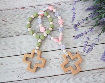 May include: Two wooden baby teething necklaces with cross-shaped pendants. One necklace features green and white beads, the other has pink and purple flower accents. The necklaces rest on a white wooden surface, with lavender sprigs in the background.