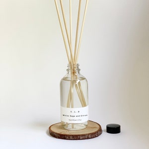 May include: A clear glass bottle with a white label that reads "C.L.O. White Sage and Citrus Reed Diffuser 14.8 oz" and a black lid. The bottle is filled with a clear liquid and has wooden sticks inserted into it. The bottle is sitting on a wooden round slice.