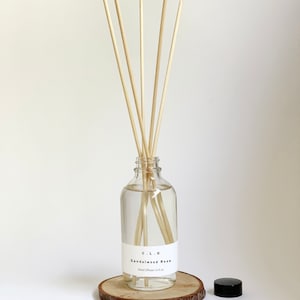 May include: A clear glass bottle with a black lid and wooden sticks, filled with a clear liquid. The bottle has a white label that reads "C.L.O. Sandalwood Rose Reed Diffuser 14.6 fl oz". The bottle is sitting on a wooden slice.